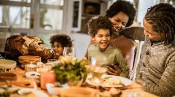 A family of four, featuring two children, sits around a Thanksgiving dinner laid out on a table, smiling.
